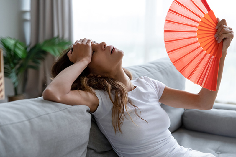 Why Is My AC Blowing Hot Air? Photo of a woman fanning herself off in her living room to stay cool.