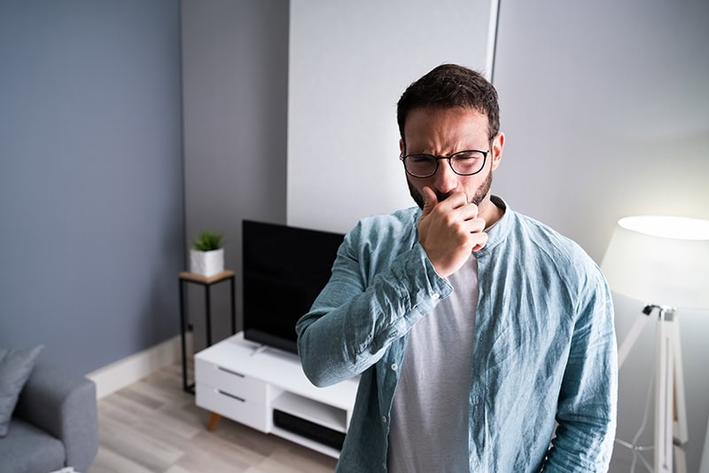 A man with a beard and glasses inside a modern living room. He is pinching his nose with his hand and grimacing with a disgusted look on his face, indicating that he is reacting to an unpleasant odor or bad smell in the air.