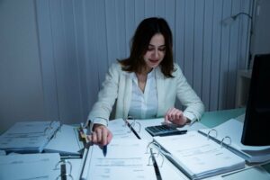 Saving Money on Your Business’s HVAC System. Image is a photograph of a woman with short brown hair and a white button down shirt sitting at a desk and reviewing paperwork.